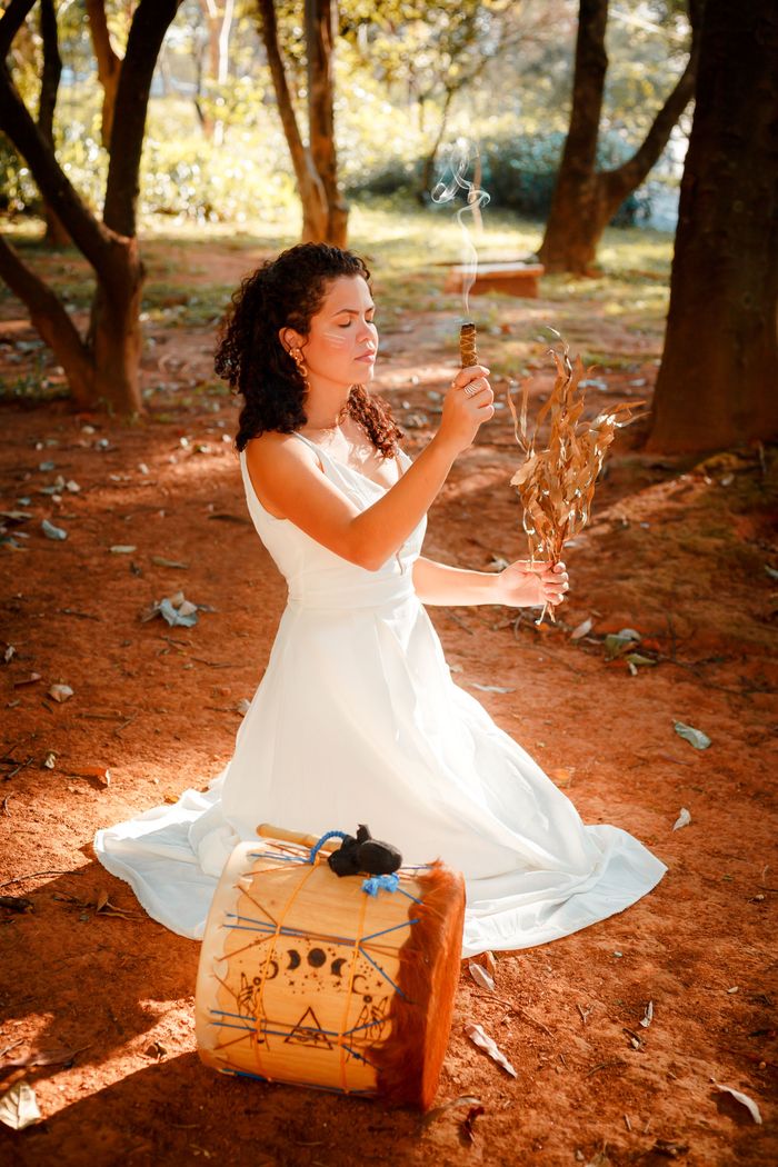brand-02 Woman in white dress meditates outdoors with drum and sage, enjoying nature's serenity.