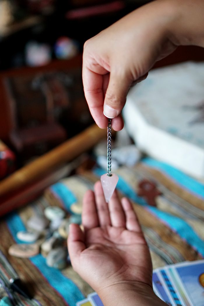 creative-01 A person holding a rose quartz pendulum over an open hand, indicating divination or energy practice.